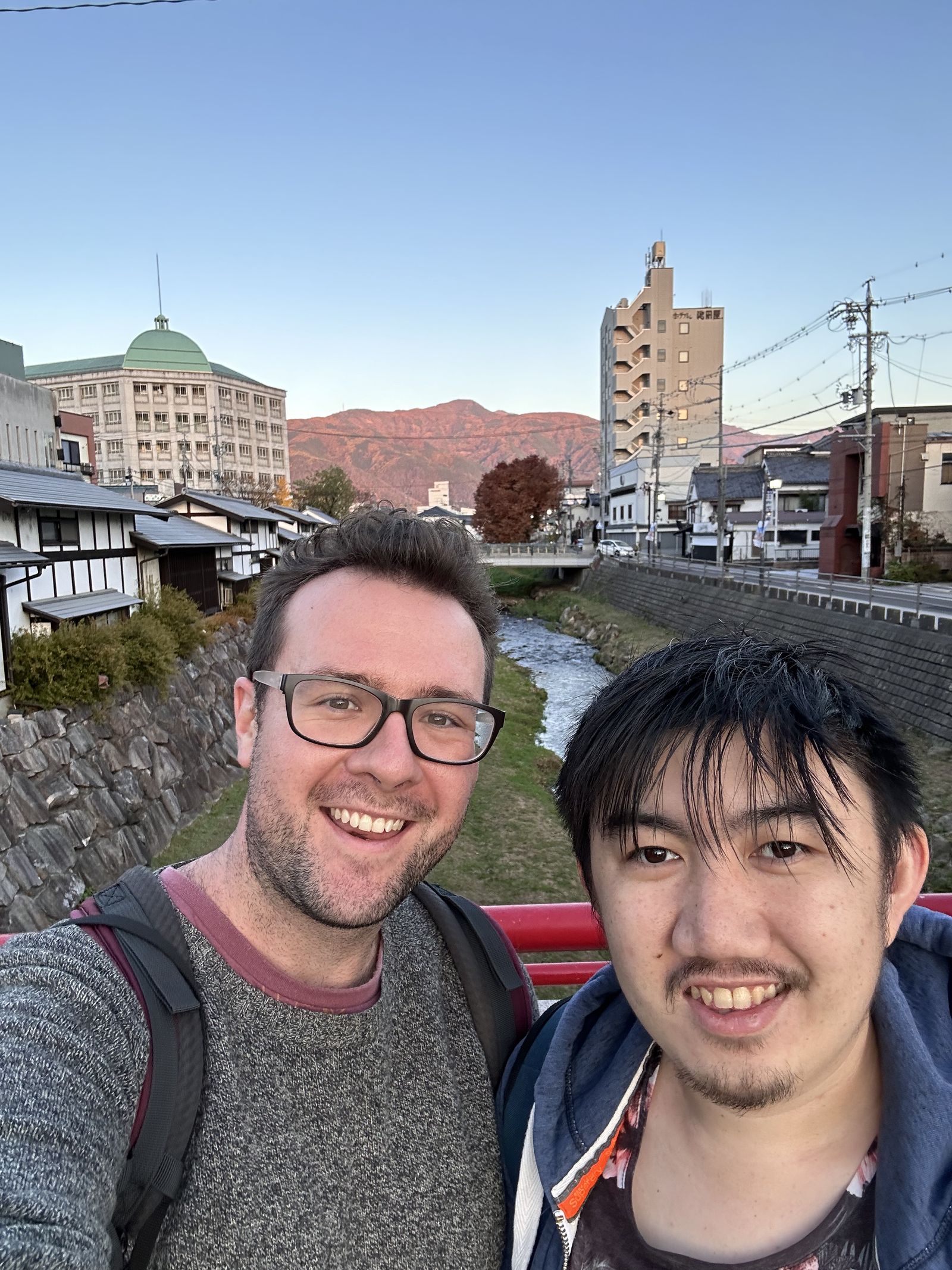 A selfie of two people smiling with a backdrop of Matsumoto city. There's a river and some mountains and some buildings on either side of the river.