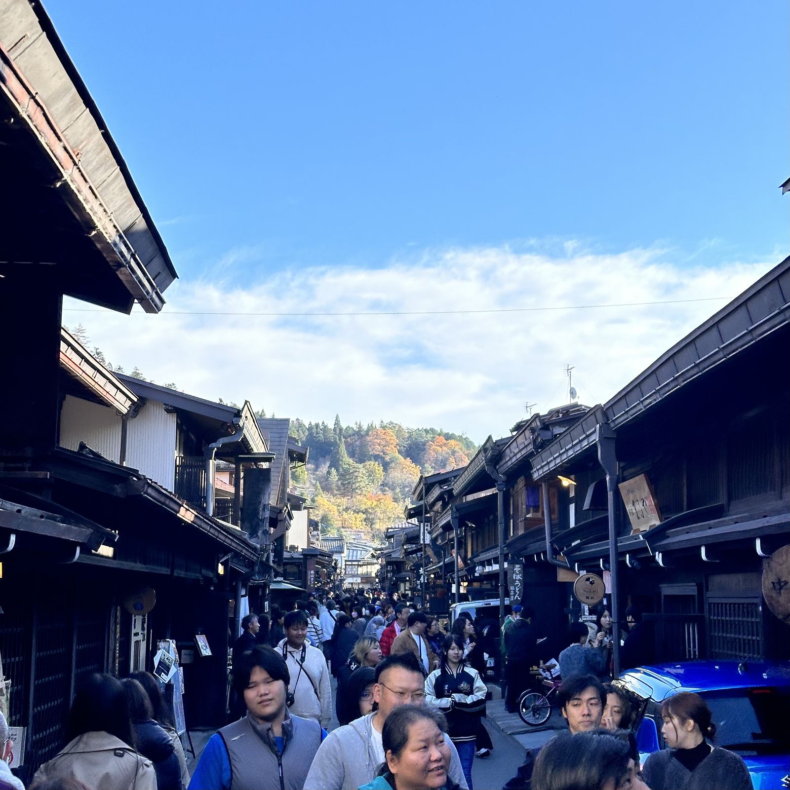 A photo of a very busy and narrow street, with many old Japanese buildings on either side, and trees in the distance