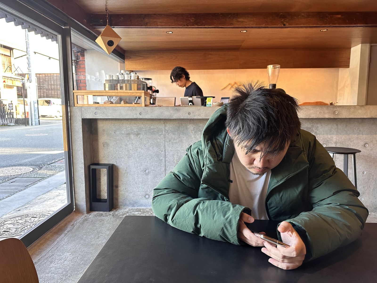 A man looking at his phone in a cafe. There is a bar with the barista at it, behind.